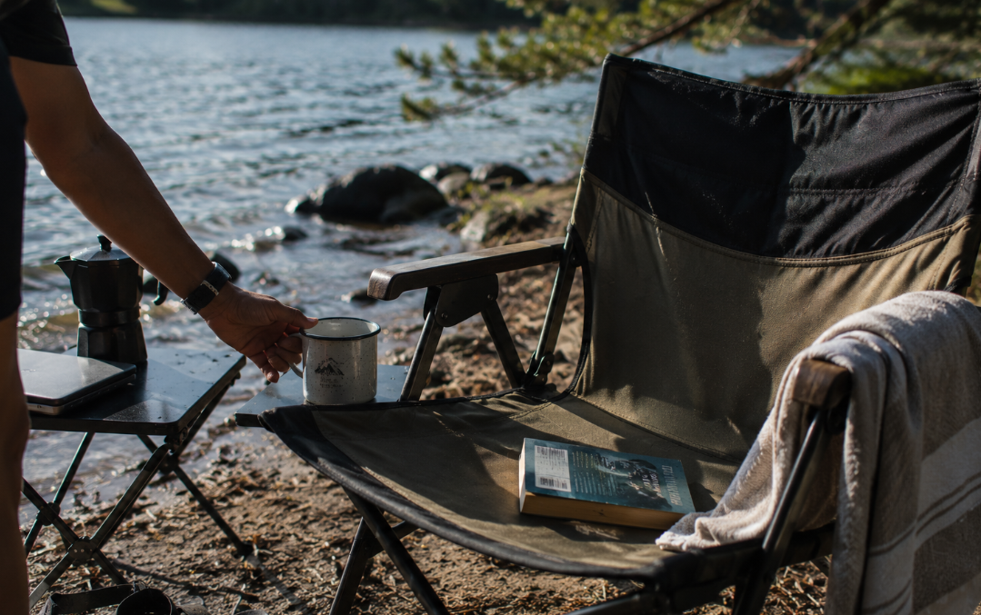 A candid lakeside afternoon scene shows a person’s hand setting a mug on a small side table beside a camping chair, with wet sandals, a towel, and rippled water nearby. The image feels relaxed and lived-in, with no visible logos or text overlay.