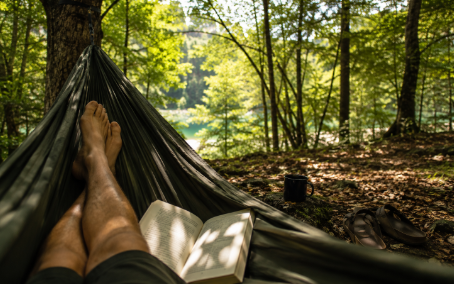 A candid daytime forest scene shows a person’s bare feet resting in a hammock, with sunlit leaves, a paperback book, and a small mug on the ground nearby. The image feels relaxed, airy, and personal, with no visible logos or text overlay.