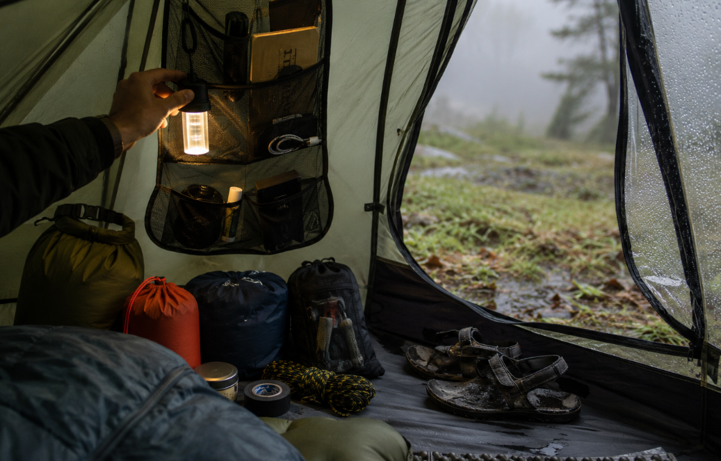 realistic early-morning tent interior scene shows a camper’s hand reaching for a hanging light, with organized pouches, dry bags, a folded mat, and muddy sandals near the open doorway. The image feels intimate, practical, and lived-in, with no visible logos or text overlay.