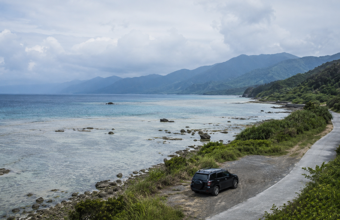 A dark SUV is parked beside a quiet coastal road with pale rocky shallows, low vegetation, and distant blue-gray mountains.