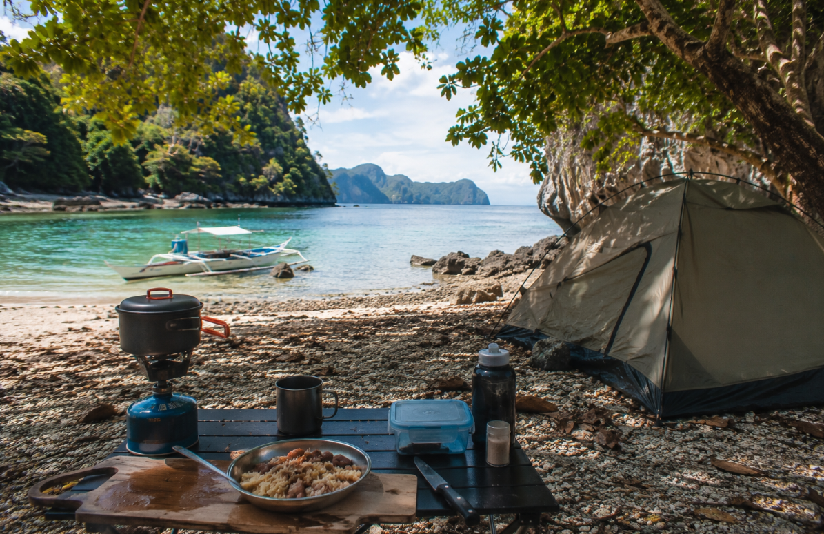 A beach campsite sits beneath leafy coastal trees, with a tent, simple cooking setup, and an outrigger boat floating in clear water near the shore.