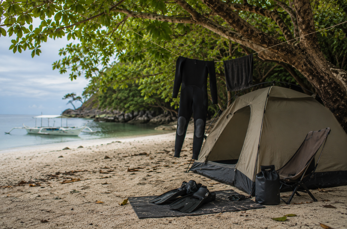 A neutral-colored tent and drying wetsuit sit beneath leafy beach trees, with dive fins, a mask, a dry bag, and a camp stool arranged on the sand near an outrigger boat.