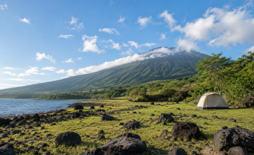 A small tent sits off to one side on grassy volcanic terrain with dark rocks in the foreground. A green volcanic slope rises in the distance under bright morning light.