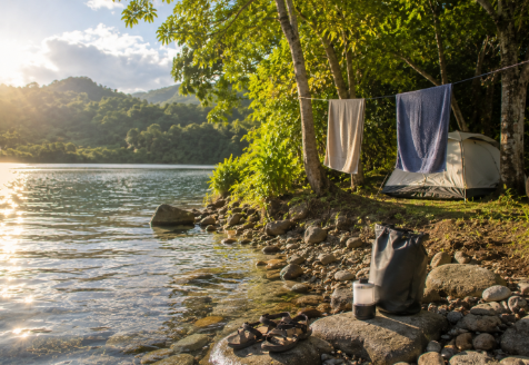 A small tent sits under trees near a calm lake, with towels drying on a line and sandals resting on smooth stones. A lantern and dry bag sit near the water as warm light reflects on the surface.